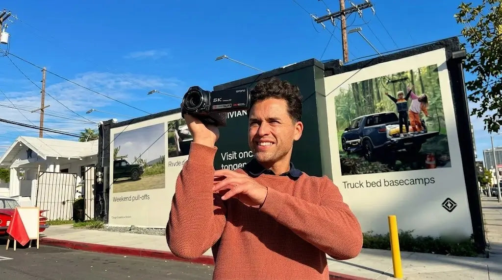A man recording with an ORDRO XV80 5K camcorder in one hand on a sunny street, demonstrating the camera size and standard outdoor operation.